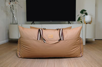 Beige travel bag with handles on a wooden floor in a room with a TV and decor.